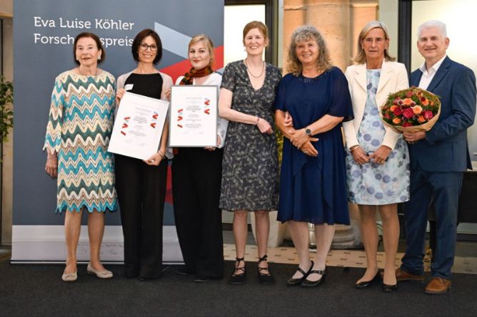 Sechs Personen stehen nebeneinander, zwei halten Urkunden, eine Person rechts hält einen Blumenstrauß, alle tragen festliche Kleidung.