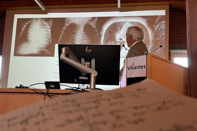 Älterer Mann steht an einem Rednerpult mit dem Schriftzug Vivantes und zeigt auf eine Projektion von Röntgenaufnahmen der Lunge hinter einem Computermonitor.