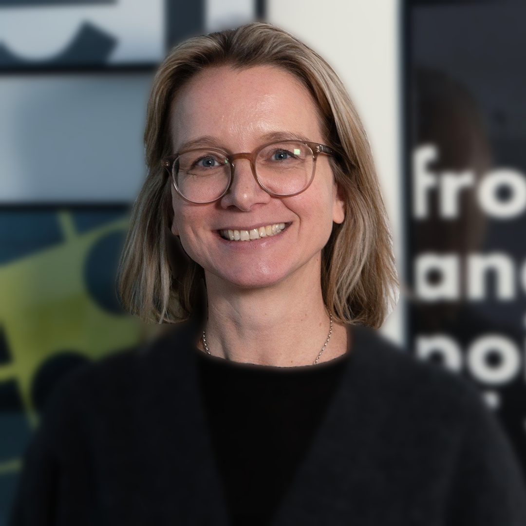 Person mit schulterlangem Haar und schwarzem Pullover vor unscharfem Hintergrund mit Textfragmenten