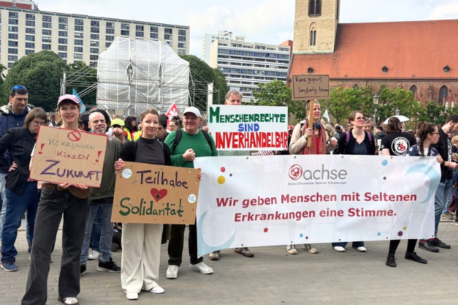 Gruppe von Menschen bei einer Demonstration mit Bannern und Schildern, darunter ein Banner mit dem Text 'Wir geben Menschen mit Seltenen Erkrankungen eine Stimme' und Schildern mit den Aufschriften 'Kürzungen Zukunft', 'Teilhabe Solidarität' und 'Menschenrechte sind unverhandelbar'