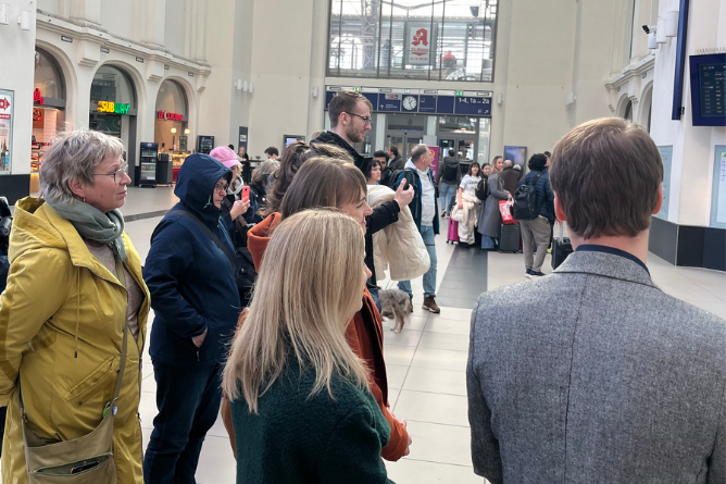 Gruppe von Menschen in einer Bahnhofshalle, im Hintergrund Geschäfte und Anzeigetafeln