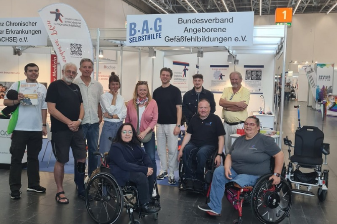 Gruppe von Menschen, darunter mehrere im Rollstuhl, vor einem Messestand mit Bannern des Bundesverbands Angeborene Gefäßerkrankungen e.V.