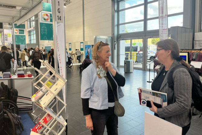 Zwei Frauen im Gespräch auf einer Messe mit Informationsständen und Publikationen im Hintergrund