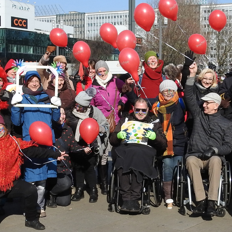 Gruppe von Menschen unterschiedlichen Alters bei einer Kundgebung im Freien. Mehrere Personen halten rote Luftballons, zwei Personen sitzen im Rollstuhl. Einige tragen Winterkleidung und bunte Gesichtsbemalung. Im Hintergrund städtische Gebäude.