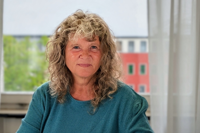 Frau mit lockigem Haar und grünem Oberteil vor Fenster mit Blick auf grüne Bäume und rotes Gebäude