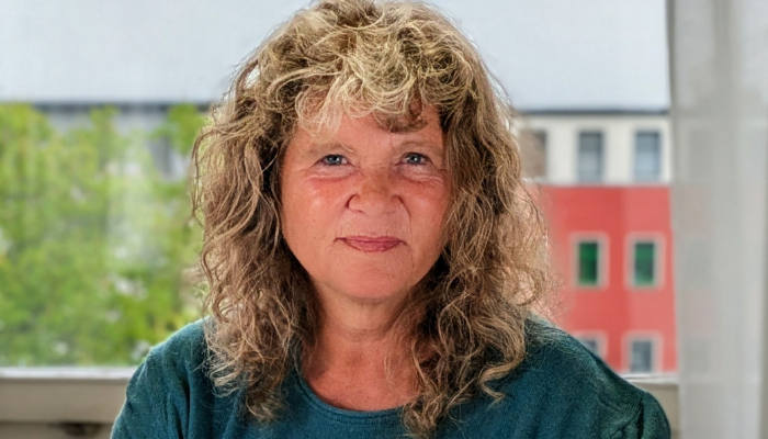 Frau mit lockigem Haar und grünem Oberteil vor Fenster mit Blick auf grüne Bäume und rotes Gebäude