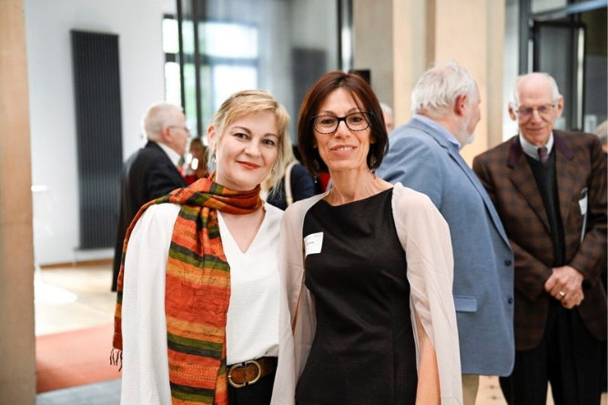 Dr. Iuso (r.) und Dr. Laugwitz Zwei Frauen stehen nebeneinander in einem hellen Raum, im Hintergrund unterhalten sich mehrere ältere Personen.