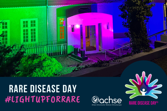 Eingang eines Gebäudes, das mit bunten Lichtern in Grün, Blau und Pink angestrahlt wird, darunter ein Banner mit dem Text 'RARE DISEASE DAY #LIGHTUPFORRARE' und dem Logo der Allianz Chronischer Seltener Erkrankungen sowie dem Logo des Rare Disease Day