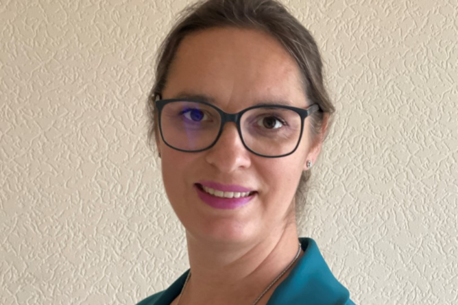 Frau mit Brille und zurückgebundenem Haar trägt ein türkisfarbenes Sakko und lächelt vor einer hellen, strukturierten Wand