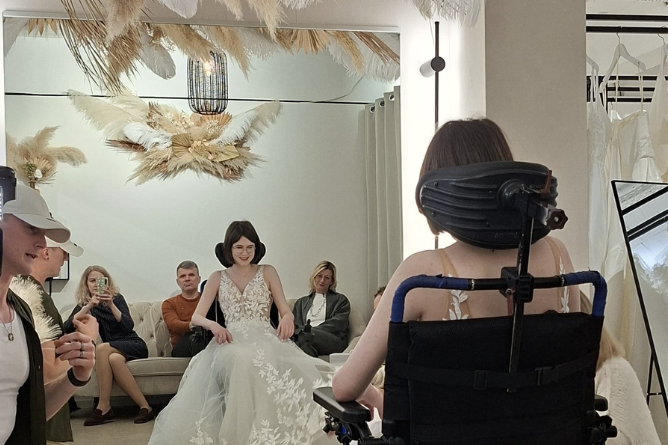 Arina im Hochzeitskleid Frau im Rollstuhl betrachtet eine Braut in einem weißen Spitzenkleid vor einem Spiegel, im Hintergrund sitzen mehrere Personen auf einem Sofa