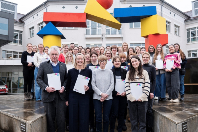 Gruppe von Menschen steht vor einem großen, bunten, geometrischen Tor aus rechteckigen und dreieckigen Formen in einem Innenhof
