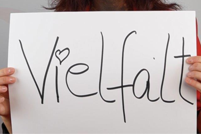 Person hält ein weißes Schild mit dem handgeschriebenen Wort 'Vielfalt' und einem kleinen Herz über dem Buchstaben 'i'