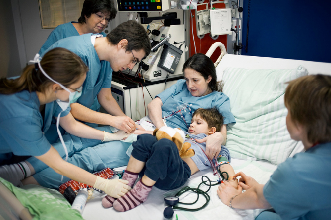 Mehrere Pflegekräfte betreuen ein Kind im Krankenhausbett, das ein Kuscheltier hält und an medizinische Geräte angeschlossen ist.