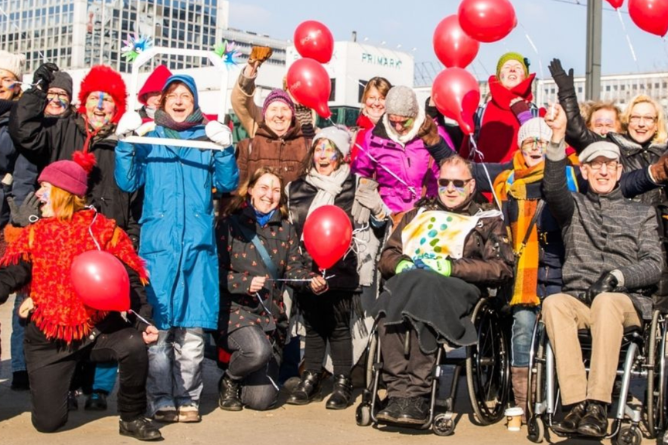 Gruppe von Menschen mit roten Luftballons, darunter eine Person im Rollstuhl, vor einem Hochhaus im Freien