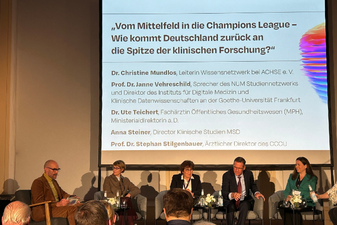 Fünf Personen sitzen auf einer Bühne vor einer Leinwand mit dem Titel 'Vom Mittelfeld in die Champions League – Wie kommt Deutschland zurück an die Spitze der klinischen Forschung' und diskutieren in einem Podiumsgespräch.