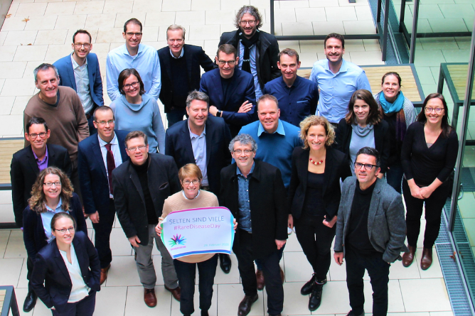 Gruppe von Menschen in Geschäftskleidung steht in einem Innenraum mit Fliesenboden, eine Person hält ein Schild mit dem Text 'SELTEN SIND VIELE #RareDiseaseDay'