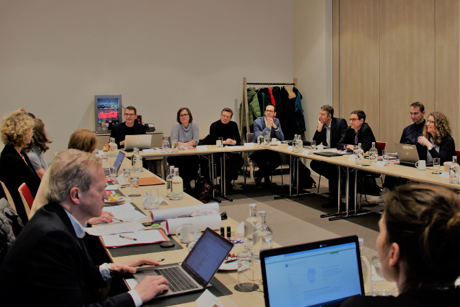 Personengruppe sitzt an einem U-förmigen Konferenztisch in einem Besprechungsraum mit Laptops und Dokumenten