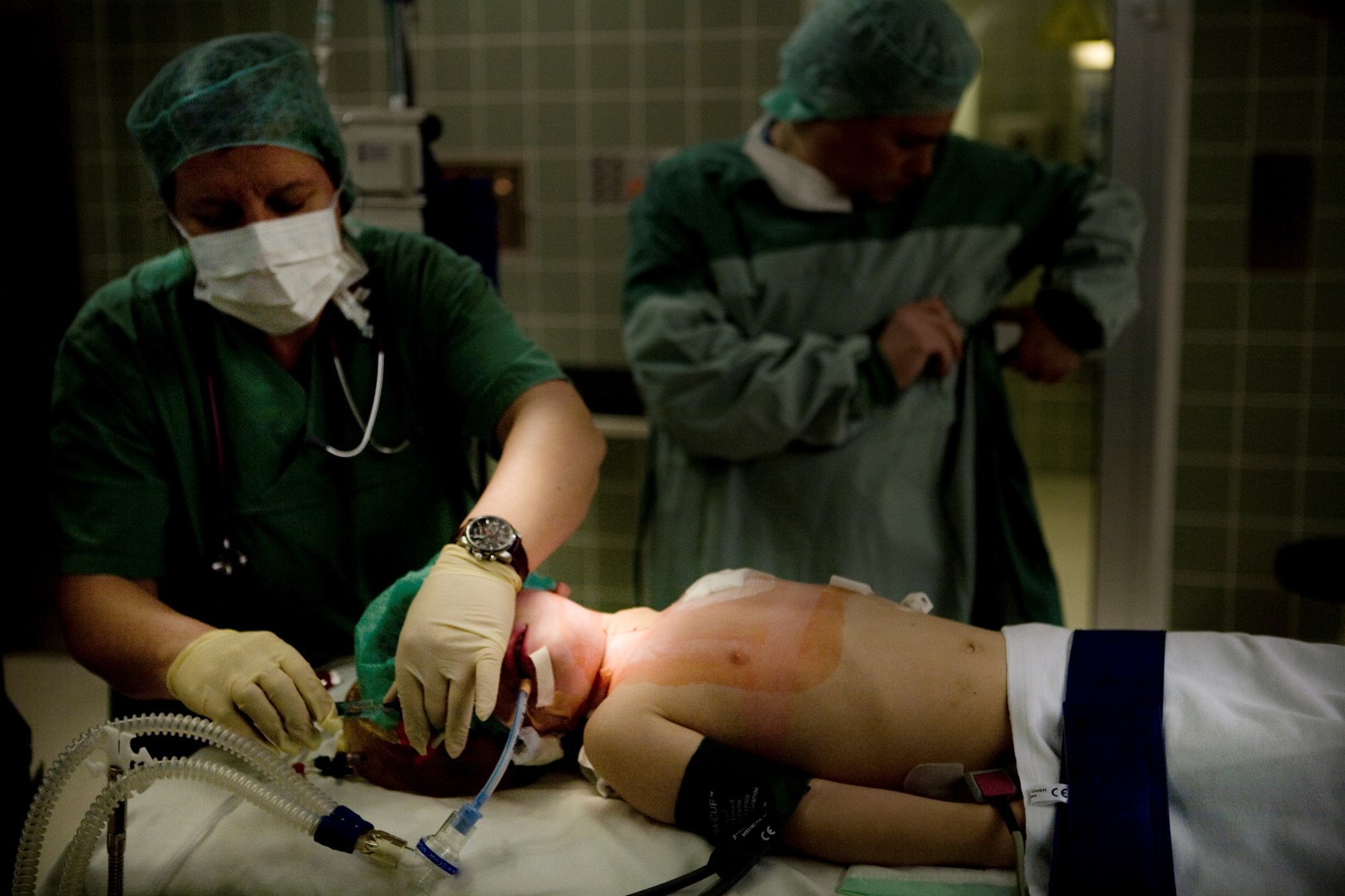Ein medizinisches Team führt einen Eingriff an einem Patienten durch, der auf einem Tisch liegt. Der Patient trägt eine Atemmaske und hat einen Zugang in den Arm. Zwei Personen in medizinischer Kleidung sind anwesend, eine Person trägt Handschuhe und eine Uhr.