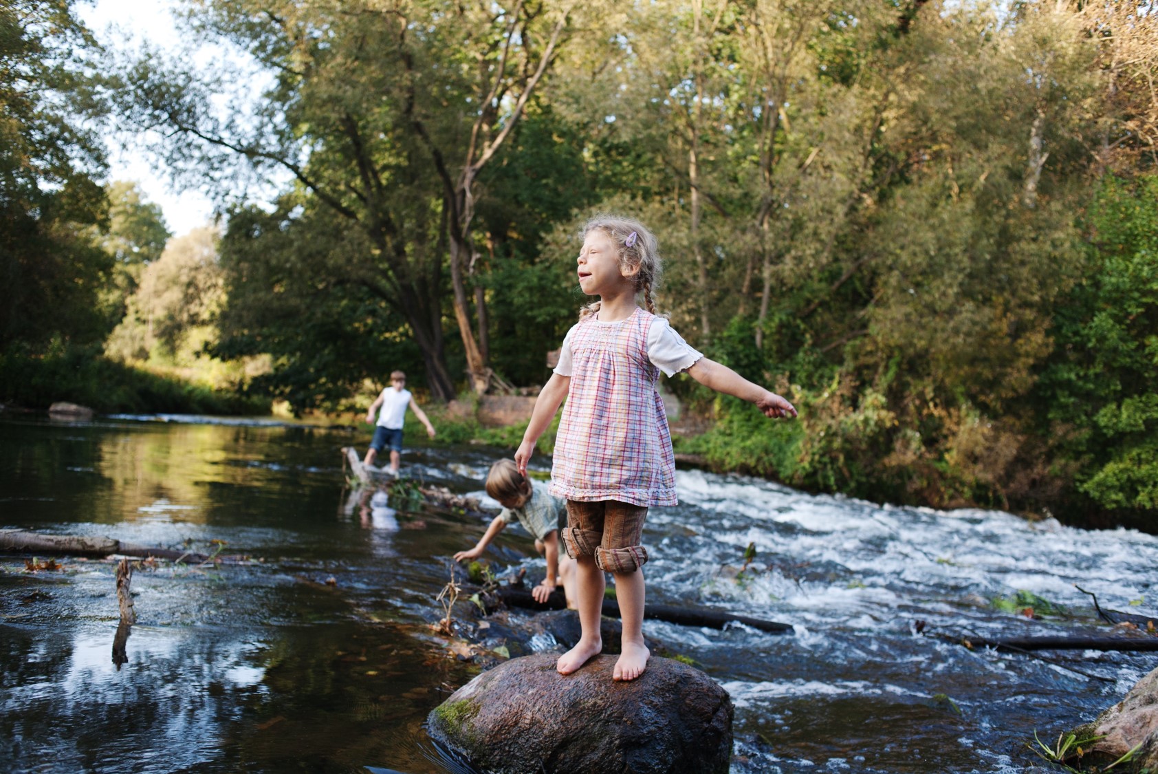 Ein Kind steht barfuß auf einem großen Stein im Wasser, trägt ein kariertes Oberteil und Shorts. Im Hintergrund spielen zwei weitere Kinder im Wasser.