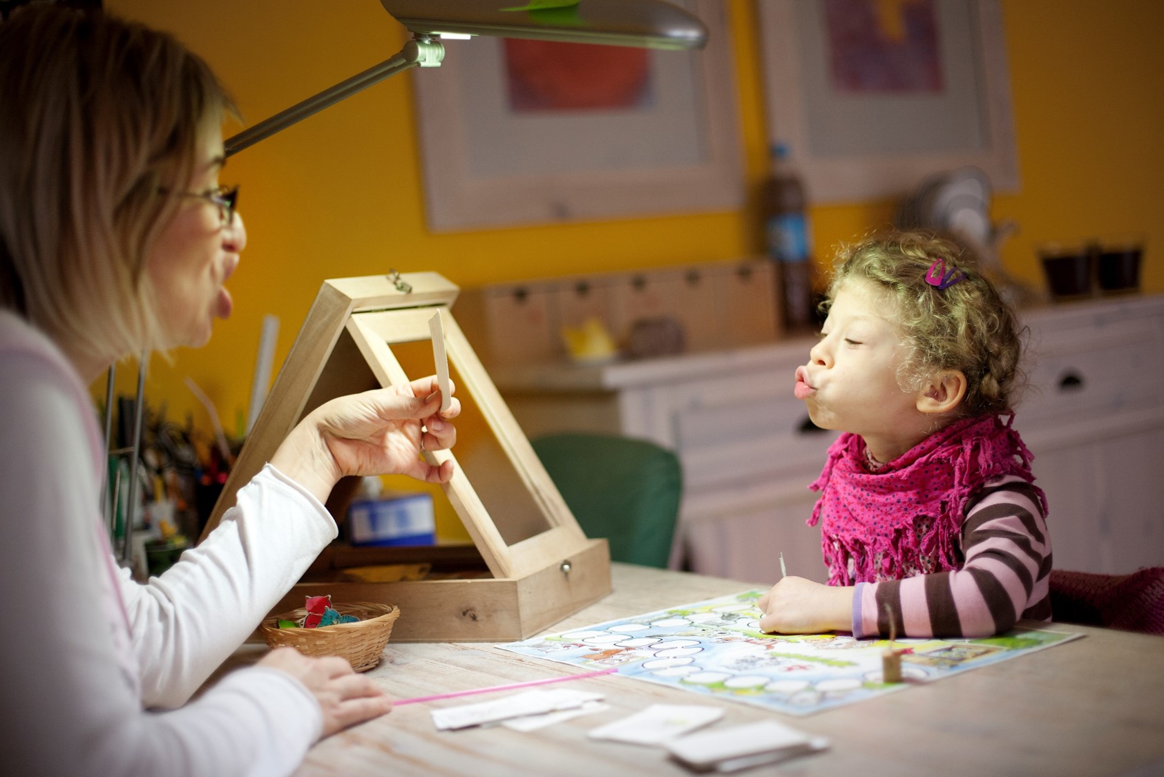 Eine Frau sitzt an einem Tisch und spricht mit einem Kind. Das Kind trägt einen rosa Schal und schaut auf ein Spielbrett mit bunten Figuren. Auf dem Tisch sind auch eine Lampe und ein Korb sichtbar.