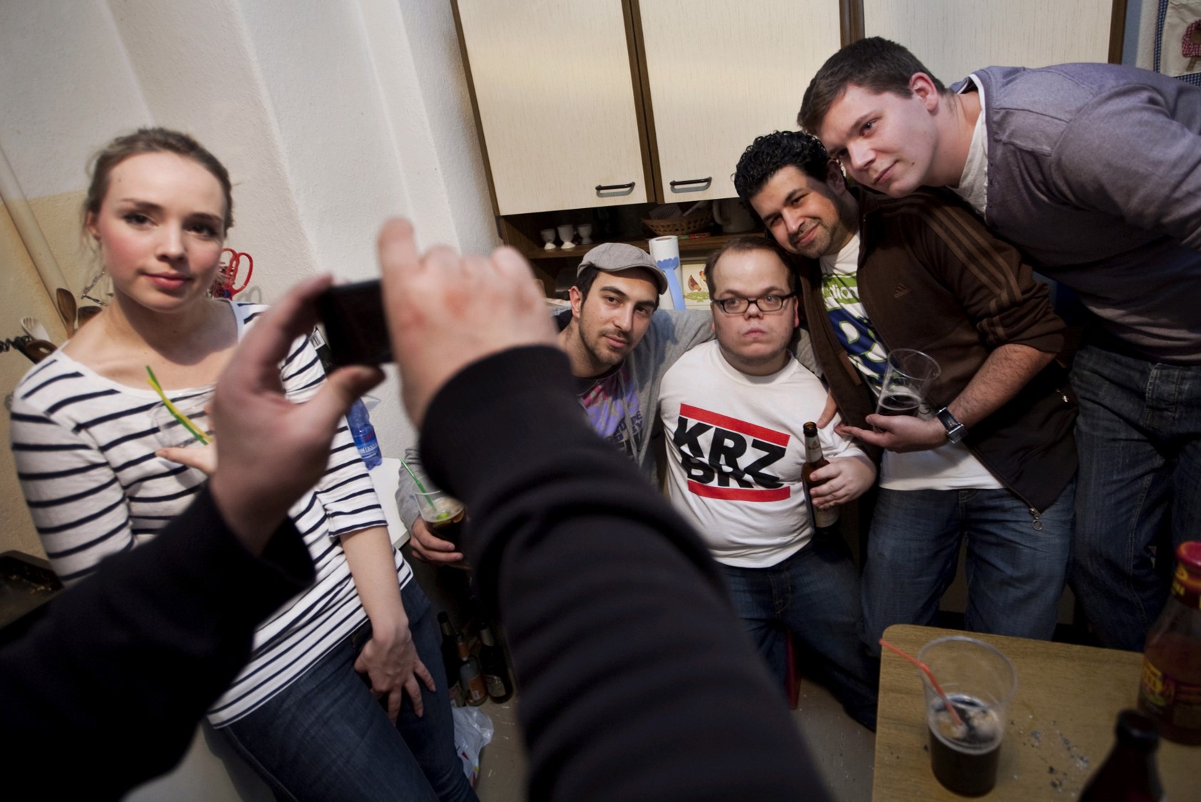 Gruppe von fünf Personen in einer Küche, eine Person hält ein Smartphone, andere haben Getränke in der Hand. Kleidung: gestreiftes Shirt, T-Shirt mit 'KRZBRG', Hemd.