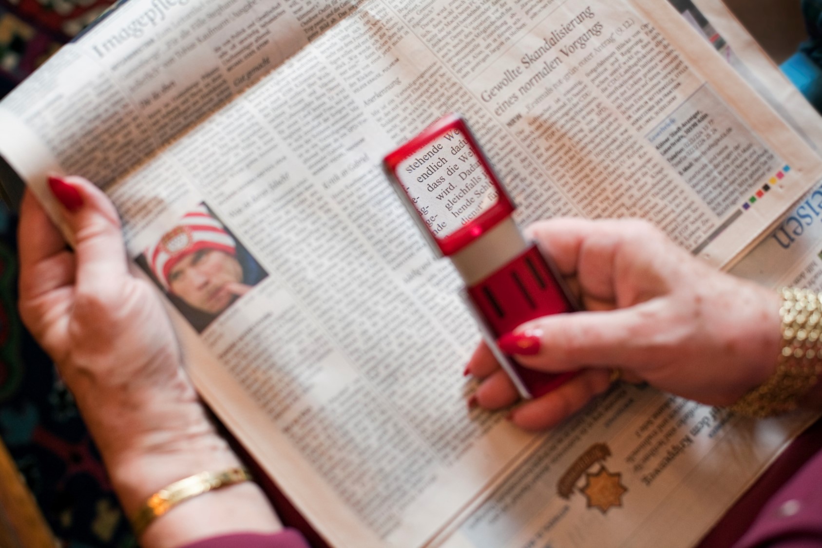 Eine Person hält eine Zeitung rot eingefasste Lupe in der Hand. Im Sichtfeld der Lupe ist der Text scharf. Außerhalb ist der Text sehr unscharf. Die Zeitung zeigt verschiedene Artikel und Bilder.