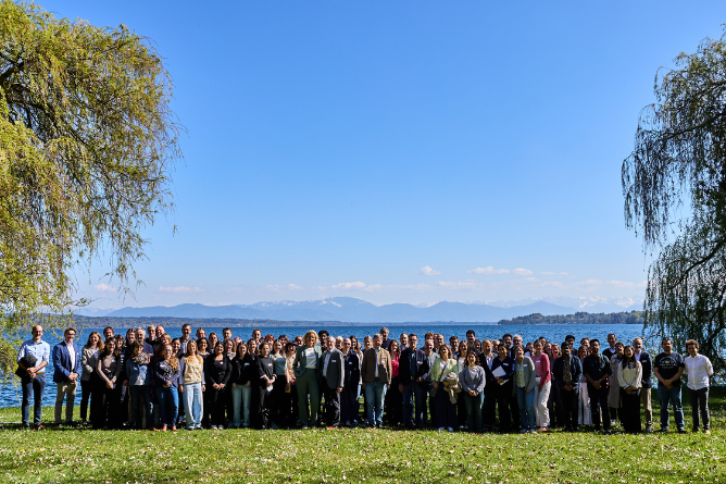Große Gruppe von Menschen steht auf einer Wiese vor einem See mit Bergen im Hintergrund und Bäumen an den Seiten