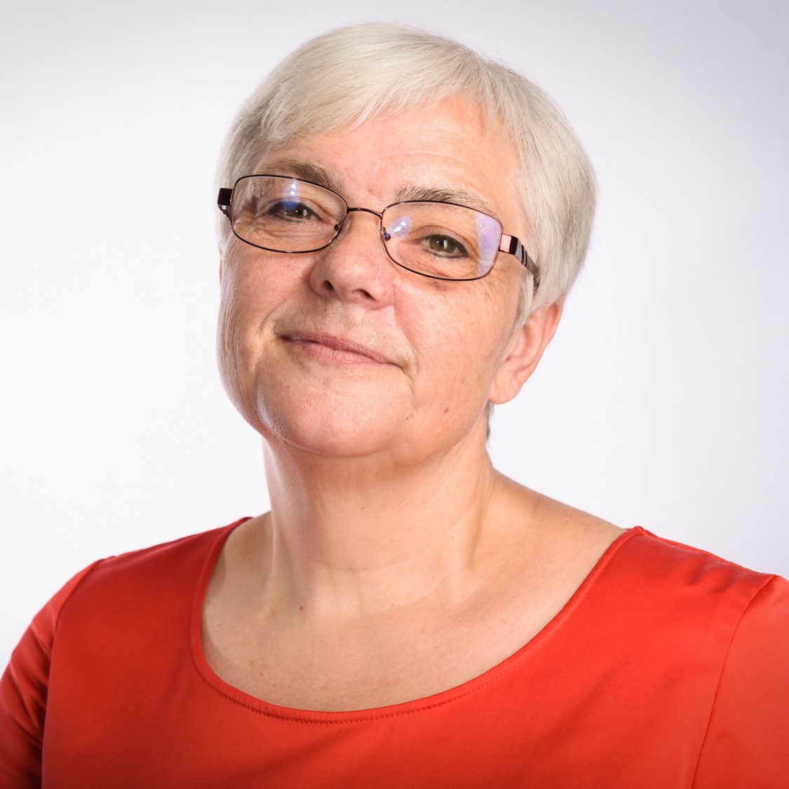 Eine Frau mit Brille, kurzen, silbergrauen Haaren. Sie trägt ein rotes Oberteil mit rundem Ausschnitt vor einem hellen, neutralen Hintergrund.