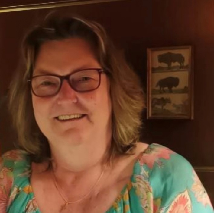 Eine Frau mit schulterlangem, hellbraunem Haar und einer Brille. Sie trägt eine sommerliche Bluse mit floralen Mustern sowie eine zarte Halskette mit einem kleinen Anhänger. Im Hintergrund eine dunkle Wand mit einem gerahmten Bild mit Tiermotiven.