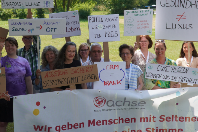 Gruppe von Menschen hält Schilder mit Botschaften zu Armut, Krankheit und Politik sowie ein Banner mit dem Schriftzug 'achse Allianz Chronischer Seltener Erkrankungen'