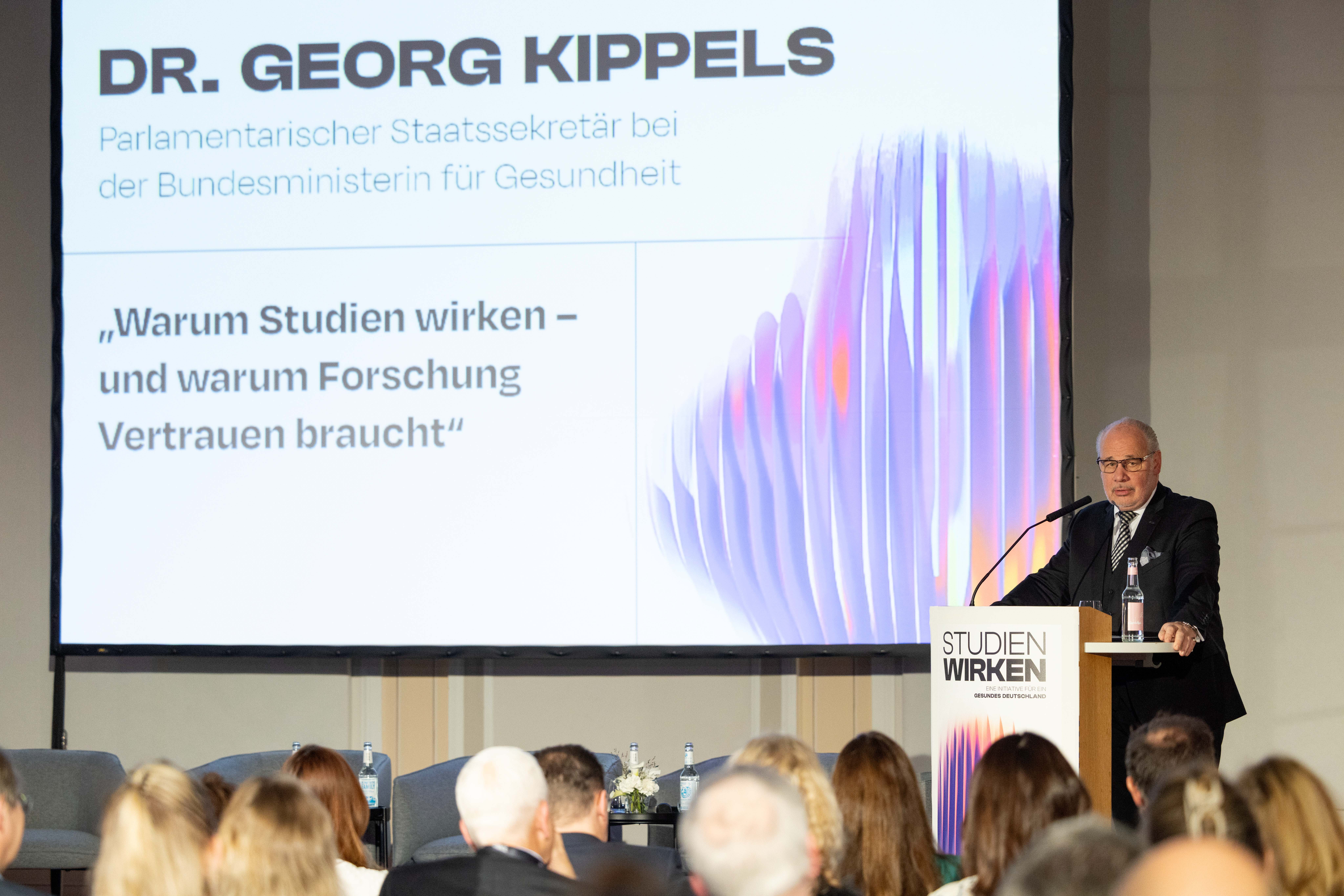 Mann steht am Rednerpult mit Mikrofon und Wasserflasche, im Hintergrund Projektion seines Vortrags, hier Vortragstitel und der Name des Vortragenden: Dr. Georg Kippels