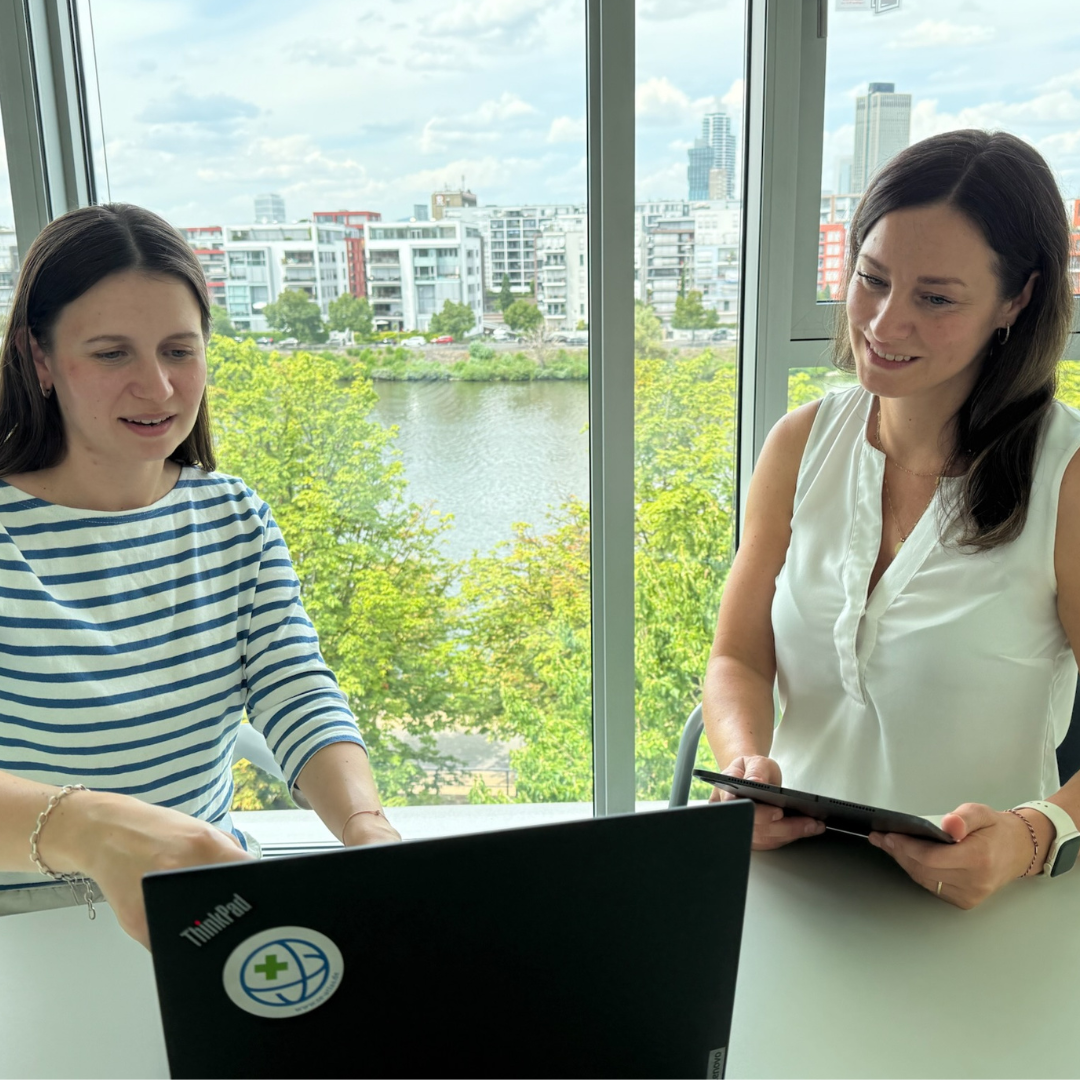 Zwei Frauen sitzen an einem Tisch vor einem Fenster mit Blick auf einen Fluss und moderne Gebäude, eine trägt ein gestreiftes Shirt, die andere ein ärmelloses weißes Oberteil, vor ihnen ein Laptop und ein Tablet