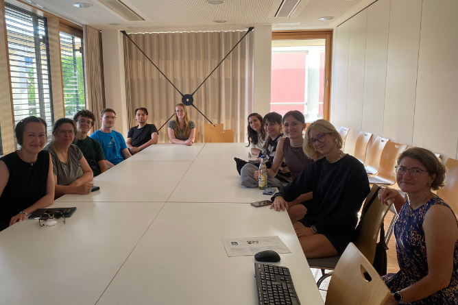 Gruppe von Studierenden sitzt an einem langen weißen Tisch in einem hellen Raum mit Fenster und Vorhang