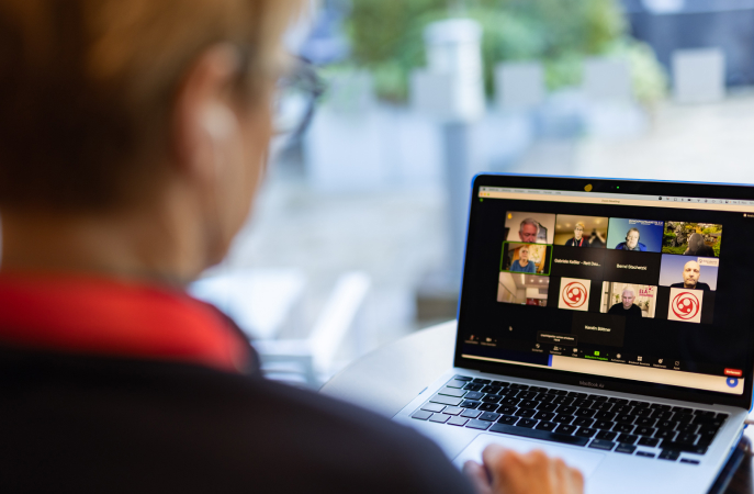 Eine Person sitzt mit dem Rücken zur Kamera und arbeitet an einem Laptop. Auf dem Bildschirm ist eine Videokonferenz mit mehreren Teilnehmenden zu sehen. Einige Teilnehmende sind sichtbar, andere werden durch das Logo der "ACHSE" (Allianz Chronischer Seltener Erkrankungen) repräsentiert. Im Hintergrund des Bildes ist ein unscharfer Außenbereich mit Pflanzen und Sitzmöglichkeiten erkennbar.