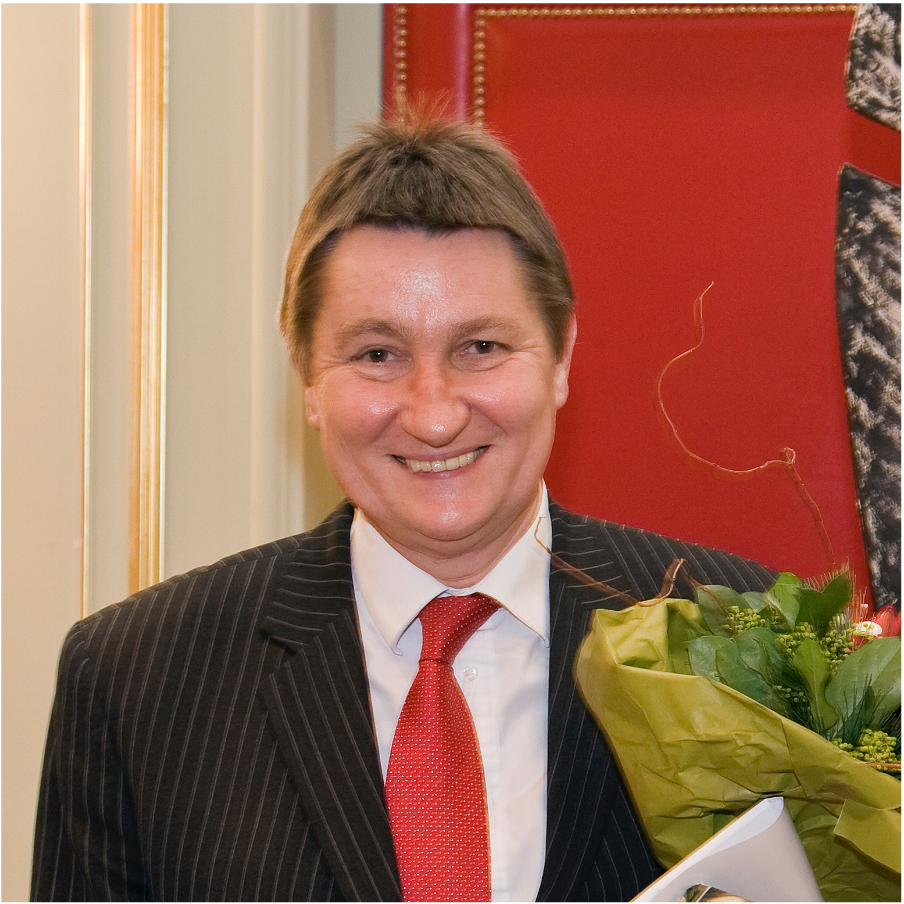 Person mit kurzem braunem Haar trägt einen schwarzen Nadelstreifenanzug, weißes Hemd und rote Krawatte, hält einen Blumenstrauß mit grünen Blättern.