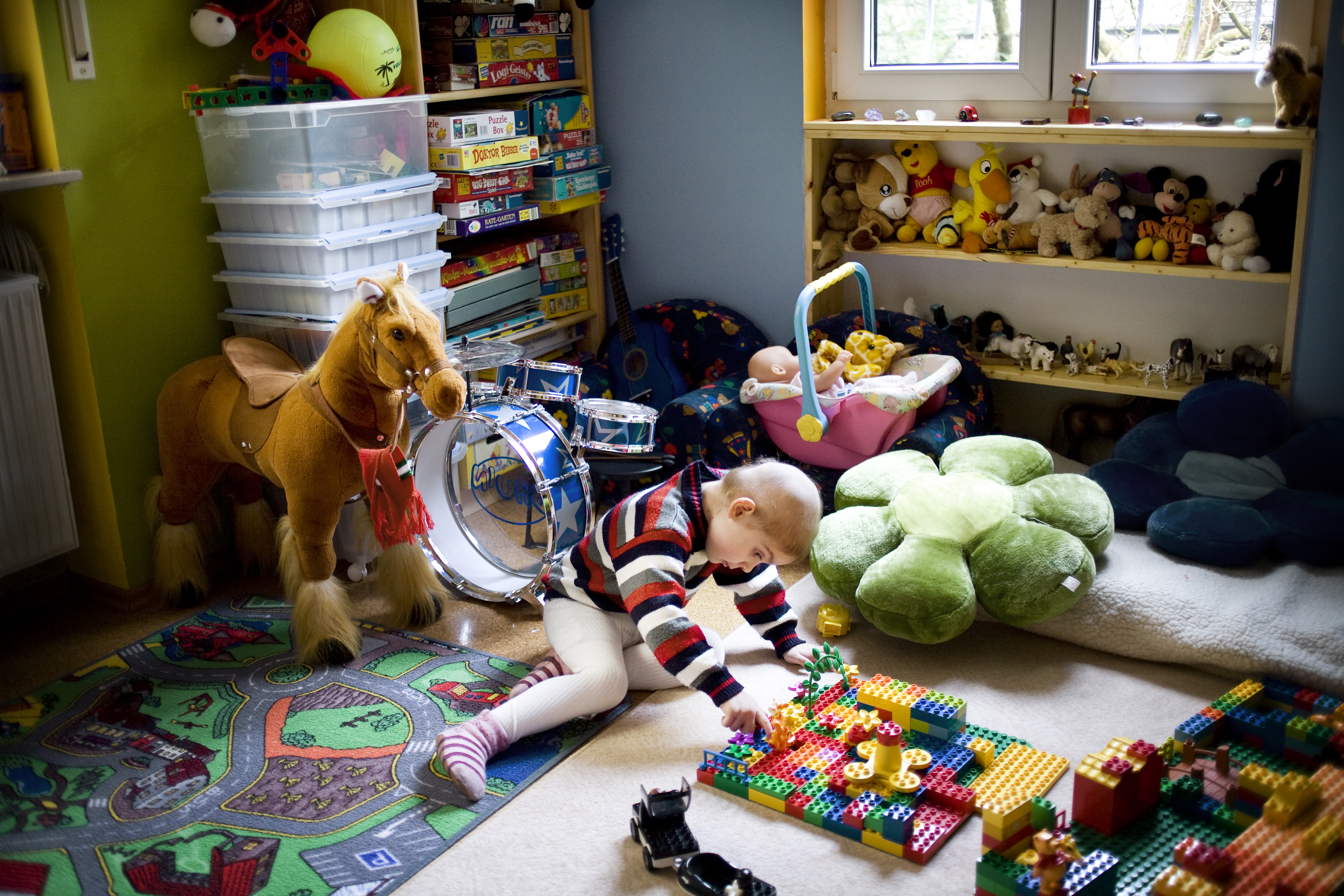 Ein Kind sitzt auf dem Boden eines Kinderzimmers und spielt mit bunten Bauklötzen. Um das Kind herum sind ein Plüschpferd, ein Spielzeugauto, eine Trommel und viele Stofftiere zu sehen. An der Wand stehen Spielzeugkisten.