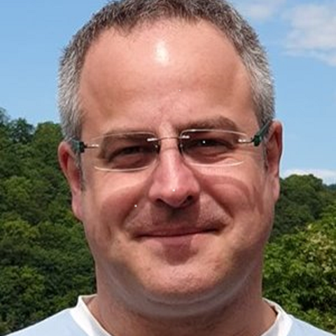 Volker Winner, ein Mann mit hoher Stirn, kurzen ergrauten Haaren, randloser schmaler Brille vor einem Hintergrund mit Wald und Himmel