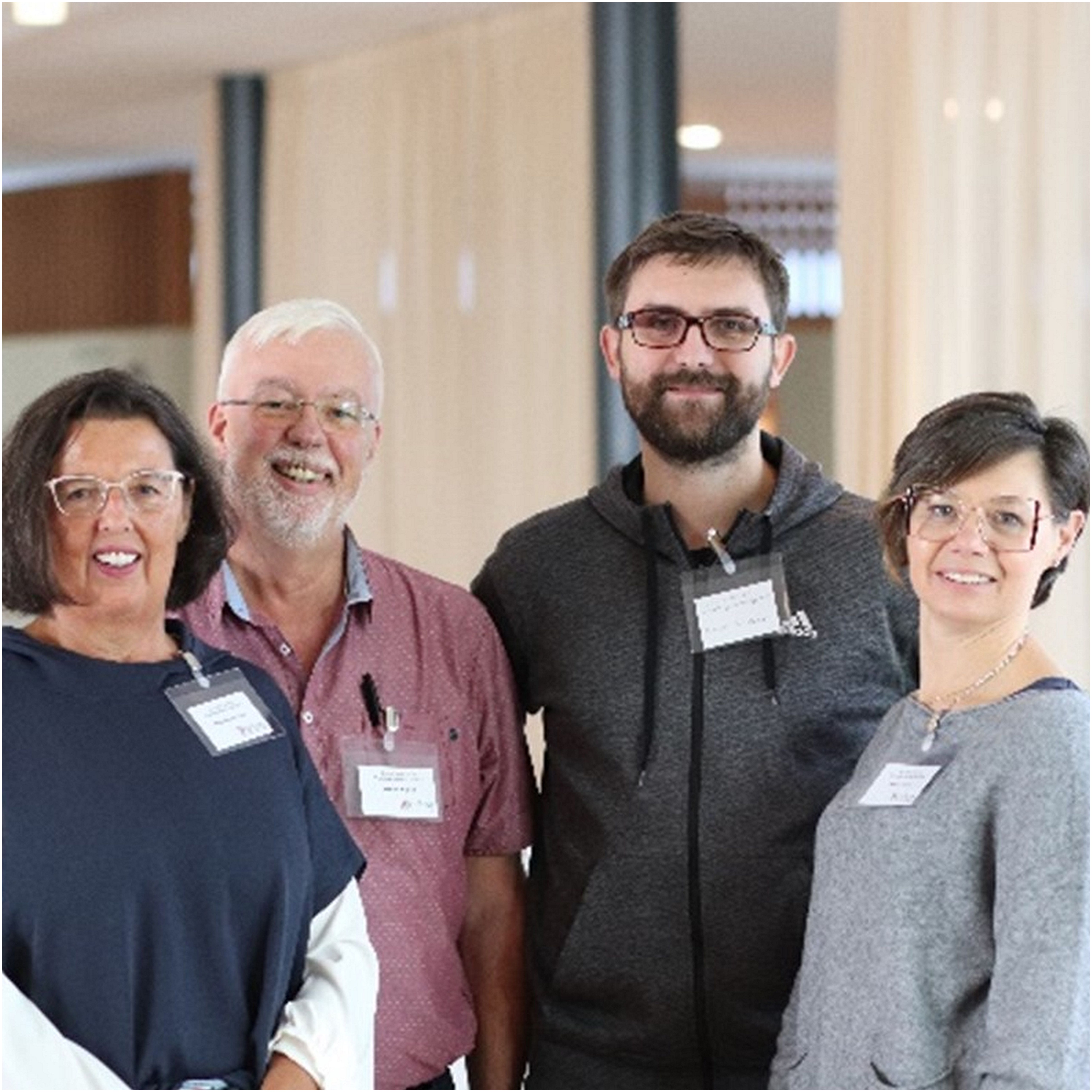 Zwei Frauen außen und zwei Männer stehen nebeneinander und lächeln in die Kamera. Sie tragen Namensschilder und befinden sich in einem hellen, modernen Innenraum mit großen Fenstern und Vorhängen im Hintergrund. Zwei Frauen außen und zwei Männer stehen nebeneinander und lächeln in die Kamera. Sie tragen Namensschilder und befinden sich in einem hellen, modernen Innenraum mit großen Fenstern und Vorhängen im Hintergrund.
