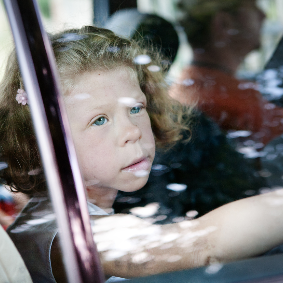 Ein junges Mädchen mit lockigem, blonden Haar sitzt in einem Auto und schaut mit nachdenklichem Blick aus dem Fenster. Ihre Hand ist an die Scheibe gelegt. Die Reflexionen auf dem Glas und die verschwommenen Personen im Hintergrund verleihen dem Bild eine melancholische Stimmung.