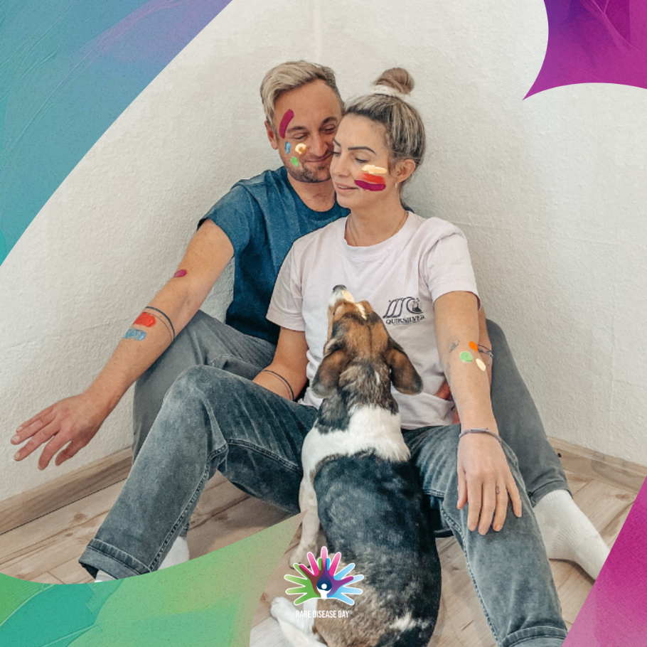 Ein Mann und eine Frau sitzen nebeneinander auf dem Boden, beide haben bunte Farben im Gesicht als Zeichen des Rare Disease Day. Vor ihnen sitzt ein Hund und schaut die Frau an. Das Bild ist von grafischen Elementen in Grün und Lila an den Ecken eingerahmt und unten ist das Logo des Rare Disease Day sichtbar.