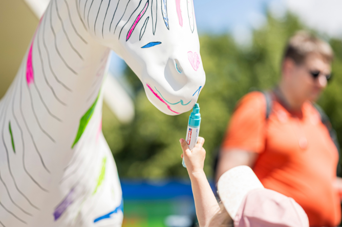 © Ravensburger Spieleland 2026 Kind malt mit blauem Stift auf ein weißes Zebra-Kunstobjekt mit bunten Markierungen im Freien