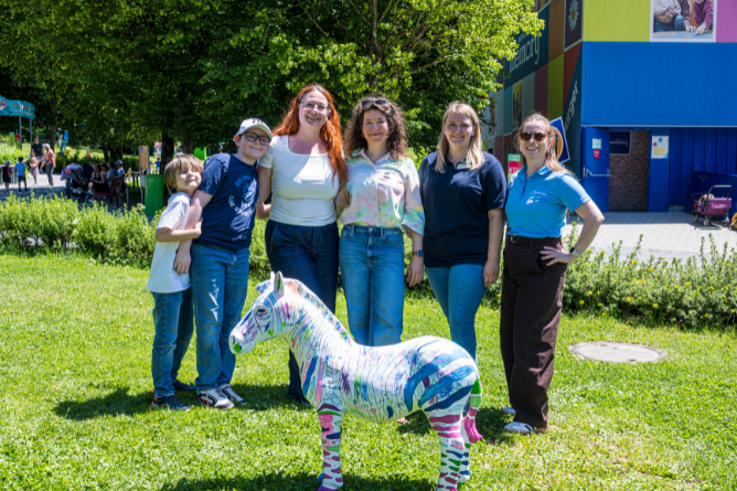 © Ravensburger Spieleland 2026 Fünf Personen stehen auf einer Wiese vor einem bunten Zebra-Skulptur, im Hintergrund Bäume und ein blaues Gebäude