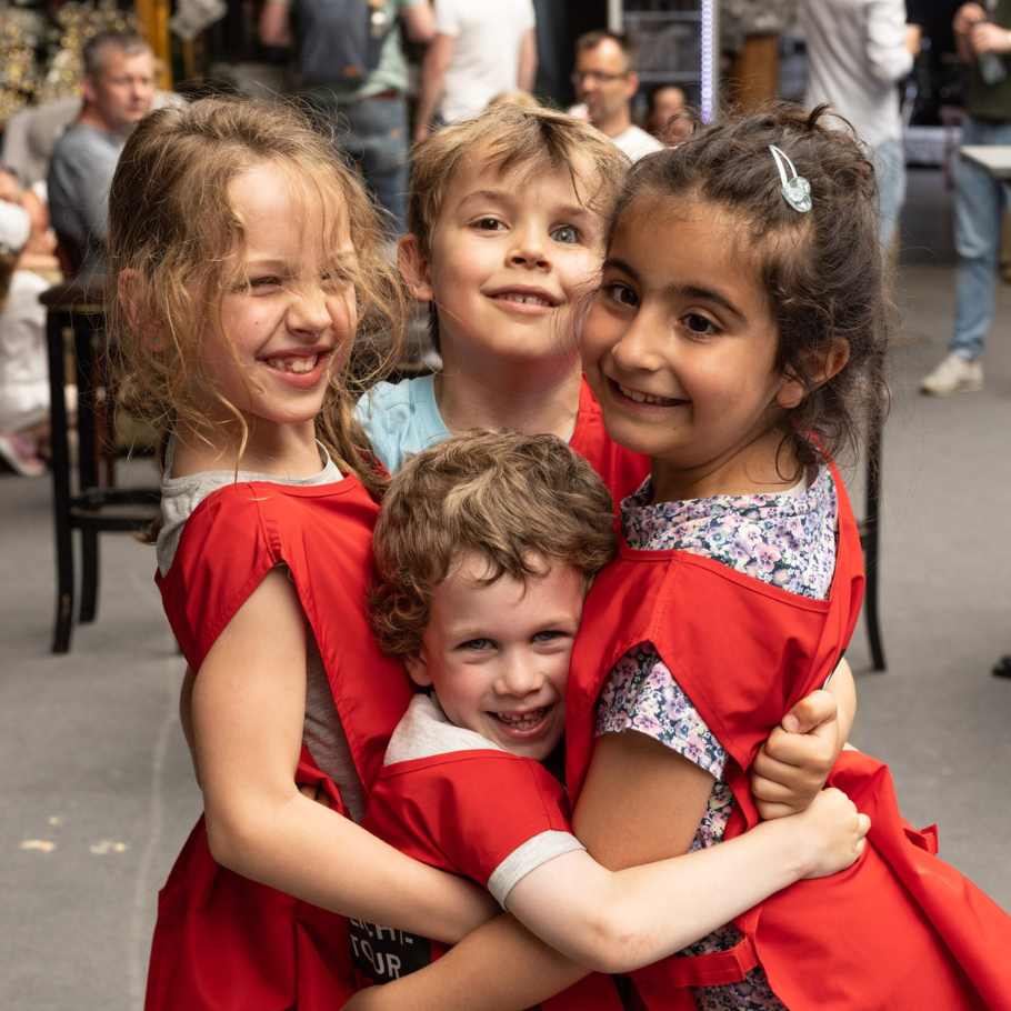 Vier lachende Kinder stehen eng umarmt in einer Gruppe zusammen. Sie tragen rote Westen, die sie als eine zusammengehörige Gruppe kennzeichnen. Im Hintergrund sind unscharf Erwachsene und Sitzgelegenheiten zu erkennen. Die Szene vermittelt Fröhlichkeit und Gemeinschaft.