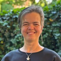 Freundlich in die Kamera schauende Frau mit zur Seite fallenden schwarz-grauen Haaren, einer Kette mit Sternen-Anhänger und einem blauen Rundhals-Sweatshirt